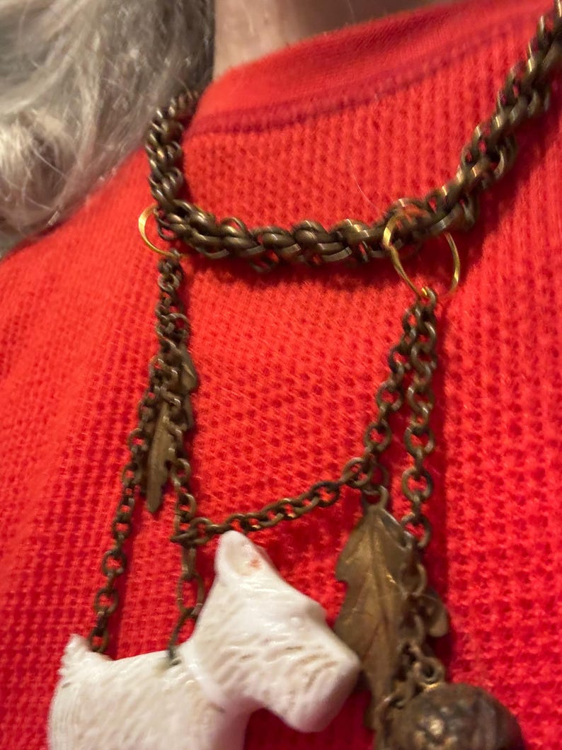 May include: A vintage necklace with a bronze chain and decorative charms. The charms include a white dog figurine, a leaf, and a small acorn. The necklace is worn over a red sweater.