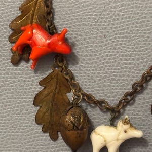 May include: A vintage necklace with woodland-themed charms. The charms include a bright orange squirrel, a white bear, a bronze oak leaf, and an acorn. The chain is a dark bronze color. The background is a light blue, textured surface.