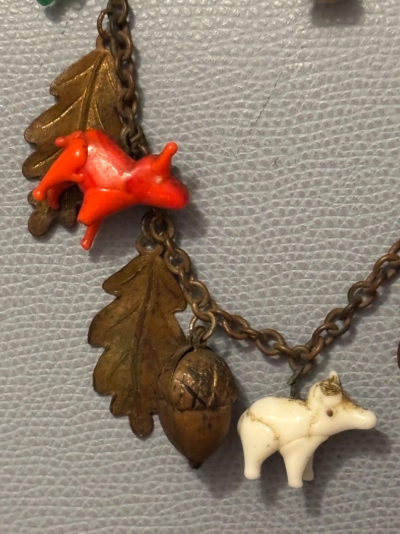 May include: A vintage necklace with woodland-themed charms. The charms include a bright orange squirrel, a white bear, a bronze oak leaf, and an acorn. The chain is a dark bronze color. The background is a light blue, textured surface.