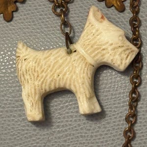 May include: Ivory-colored pendant in the shape of a Scottish Terrier dog, with etched fur details and a white collar. The dog is suspended from a bronze-toned chain with decorative oak leaf accents. A charming, vintage-style necklace.