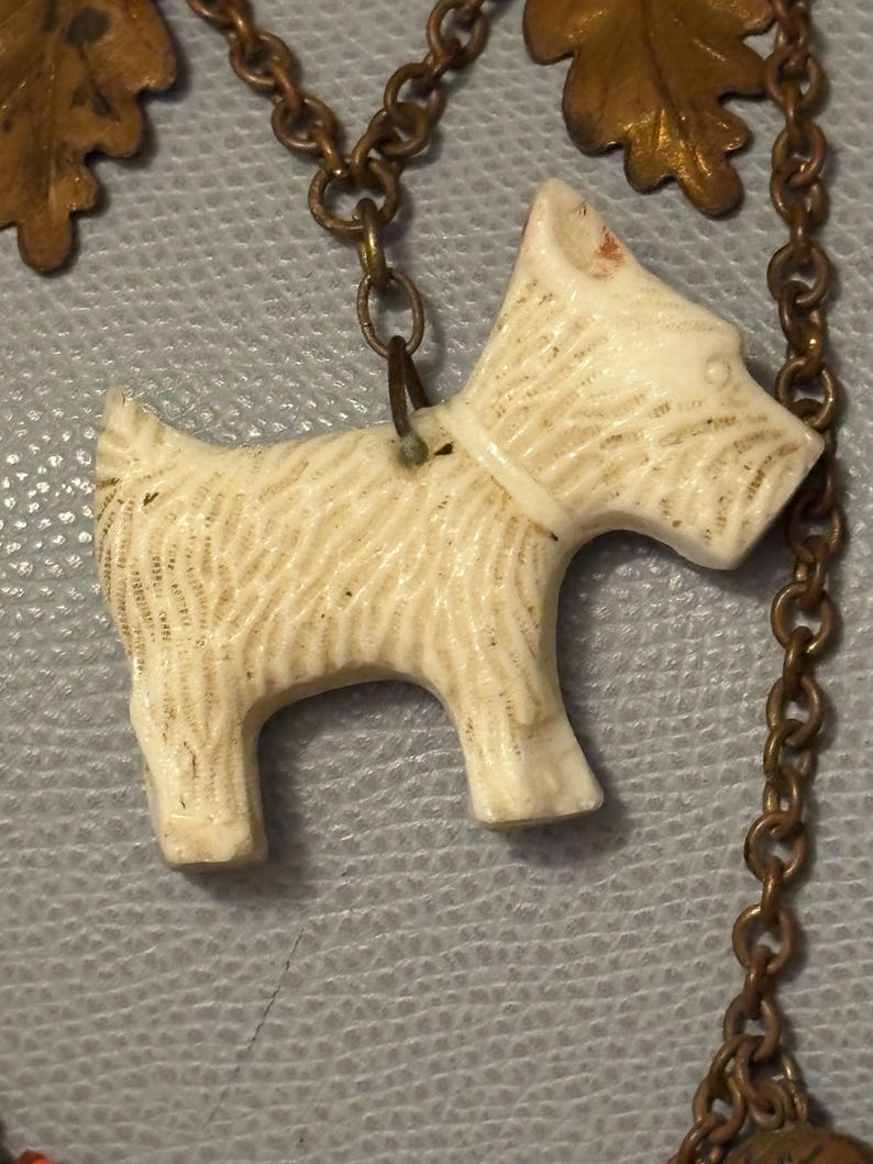 May include: Ivory-colored pendant in the shape of a Scottish Terrier dog, with etched fur details and a white collar. The dog is suspended from a bronze-toned chain with decorative oak leaf accents. A charming, vintage-style necklace.