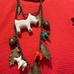 An antique 1930s charm necklace with a bronze chain and various glass molded shapes charms. The charms include white terrier dogs, green and orange animals, bronze oak leaves, and acorns. The necklace is displayed against a red background.