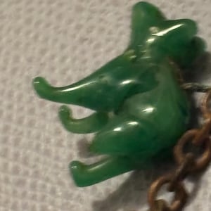 May include: A small, green, plastic elephant charm with a detailed trunk and legs. The elephant is attached to a bronze-colored chain. The charm has a glossy finish and is set against a textured, white background.