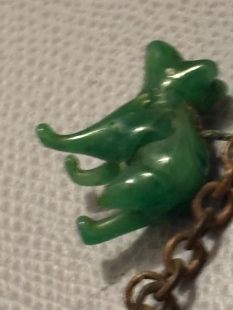 May include: A small, green, plastic elephant charm with a detailed trunk and legs. The elephant is attached to a bronze-colored chain. The charm has a glossy finish and is set against a textured, white background.