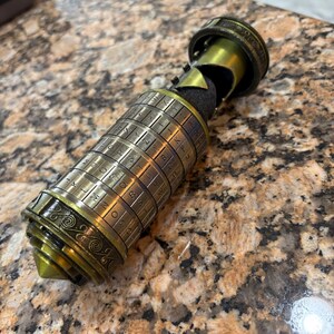 May include: A brass-colored cylindrical puzzle with rotating rings of letters. The puzzle has a conical tip and a decorative top, with a section that appears to be open. The object is on a speckled brown surface.