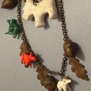 May include: A vintage necklace with a bronze chain and various charms. The charms include white dog figurines, acorns, oak leaves, a green frog, and a red squirrel. The necklace is displayed against a light blue background.
