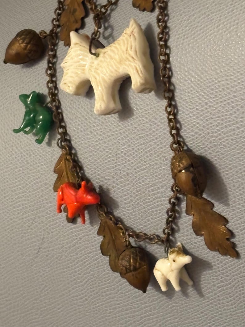 May include: A vintage necklace with a bronze chain and various charms. The charms include white dog figurines, acorns, oak leaves, a green frog, and a red squirrel. The necklace is displayed against a light blue background.