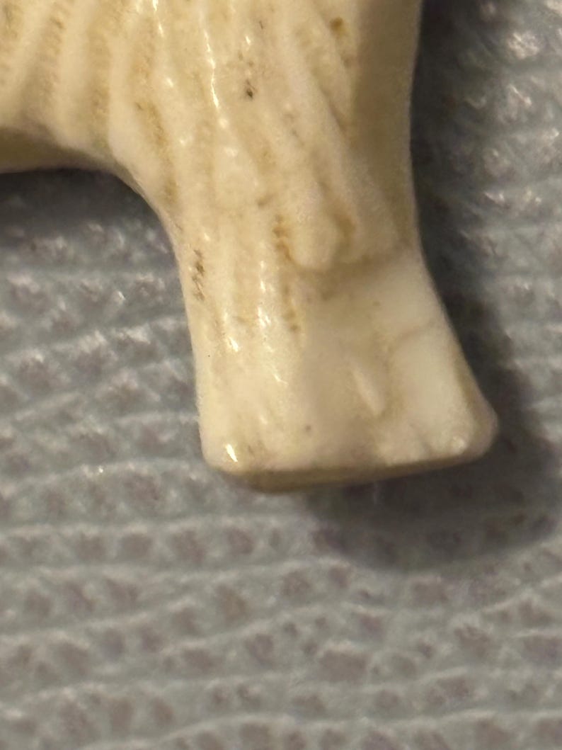 May include: Close-up of a cream-colored ceramic figurine's leg. The leg has a smooth, glossy finish with subtle brown markings and a rounded foot. The figurine appears to be a small animal or creature.