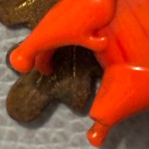 May include: Close-up of a bright orange plastic object with rounded edges, possibly a toy or decorative element. The object is set against a blurred background, with a portion of a brown, leaf-shaped object visible.