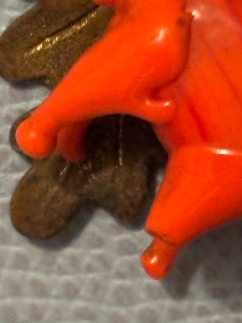 May include: Close-up of a bright orange plastic object with rounded edges, possibly a toy or decorative element. The object is set against a blurred background, with a portion of a brown, leaf-shaped object visible.