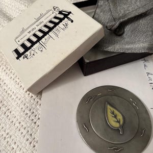 May include: A vintage metal leaf-shaped pin with a yellow center, resting on a white surface. The pin is in a box with the text "Trading Willy Nilly Company" and a black and white illustration. A gray pouch is also present.