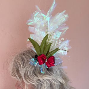 May include: A decorative headpiece featuring a cluster of white, iridescent, and green elements. A red bow with a silver gem is centered on the headpiece. The headpiece is adorned with green leaves and white flower buds.