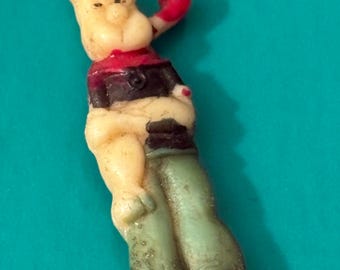May include: A vintage figurine of a cartoon character in a boxing pose. The figure has a cream-colored body, orange hair, a red boxing glove, and green pants. The figure is on a teal background.