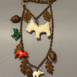 May include: A vintage-style necklace with a gold-tone chain. The necklace features various charms, including a white terrier, a green frog, a red fox, acorns, and oak leaves. The charms are suspended from the chain by smaller chains.
