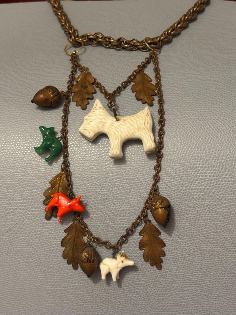 May include: A vintage-style necklace with a gold-tone chain. The necklace features various charms, including a white terrier, a green frog, a red fox, acorns, and oak leaves. The charms are suspended from the chain by smaller chains.