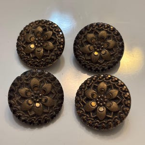 May include: Four antique gold-colored buttons with a raised floral design. The buttons have a dark brown rim and a raised center with a flower design.