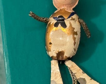 May include: Vintage sailor doll figurine with a white hat, blue collar, and gold accents. The doll has a painted face with red lips and is made of metal with visible wear and tear. The legs and arms are connected with silver chain.