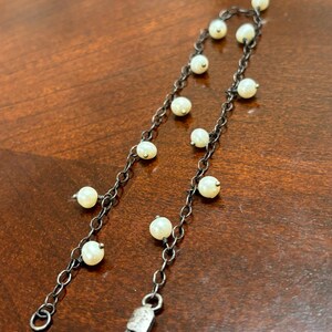 May include: A delicate silver chain bracelet featuring small, round, off-white pearls. The bracelet has a lobster clasp closure and is displayed on a wooden surface. The pearls are evenly spaced along the chain, creating an elegant and understated design.
