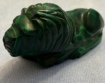 May include: A green malachite lion sculpture. The lion is lying down with its head turned to the side. The sculpture is carved with intricate details and has a smooth, polished finish.