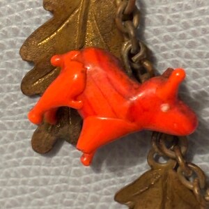 May include: Close-up of a vintage necklace featuring a bright orange plastic horse charm. The horse is connected to a brass chain with brass oak leaf charms. The chain is linked together with small brass links. The background is a light blue.