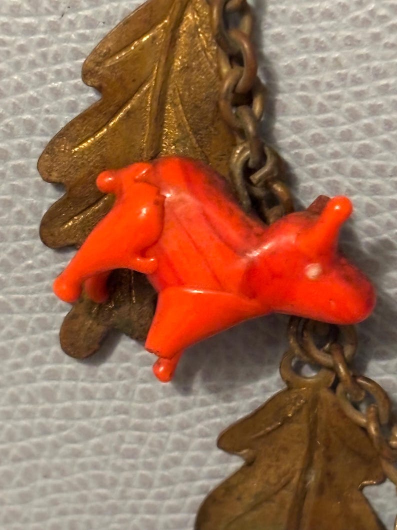 May include: Close-up of a vintage necklace featuring a bright orange plastic horse charm. The horse is connected to a brass chain with brass oak leaf charms. The chain is linked together with small brass links. The background is a light blue.