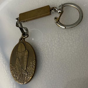 May include: A gold-toned keychain featuring a detailed depiction of a space shuttle taking off. The keychain has a simple, rectangular bar and a silver ring.