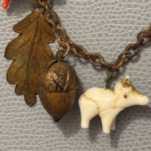 May include: A vintage charm bracelet with nature-themed charms. The charms include a bronze oak leaf, an acorn, and a small white animal figure. The bracelet has a bronze chain and a red plastic charm.