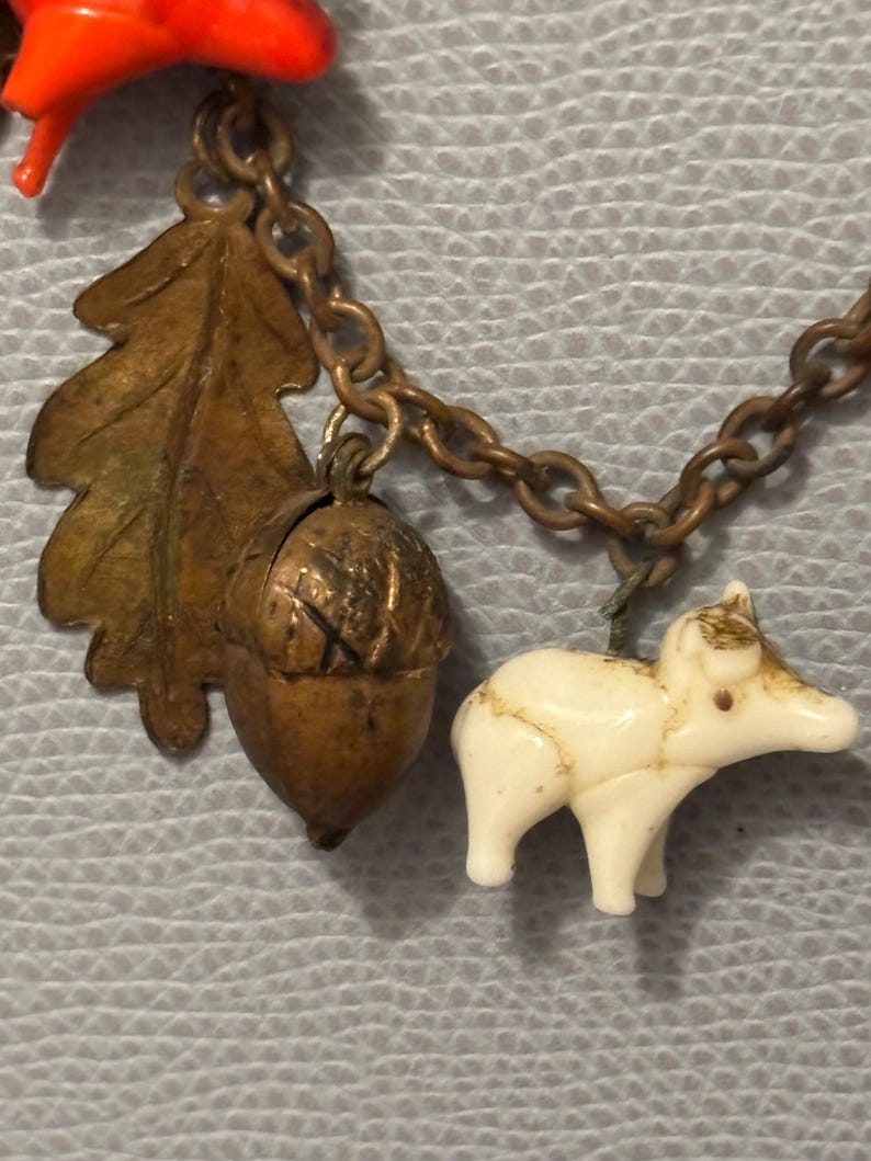 May include: A vintage charm bracelet with nature-themed charms. The charms include a bronze oak leaf, an acorn, and a small white animal figure. The bracelet has a bronze chain and a red plastic charm.