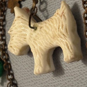 May include: Close-up of a cream-colored dog pendant with textured fur detail, attached to a bronze-toned chain. The pendant is a stylized representation of a terrier breed. The chain also features decorative leaf charms.
