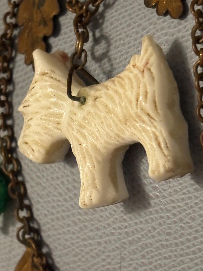 May include: Close-up of a cream-colored dog pendant with textured fur detail, attached to a bronze-toned chain. The pendant is a stylized representation of a terrier breed. The chain also features decorative leaf charms.