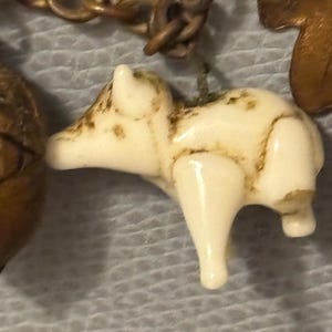 May include: A small, off-white pig charm with brown accents hangs from a bronze-colored chain. The pig is detailed with visible legs and a rounded body. The charm is part of a larger piece of jewelry.
