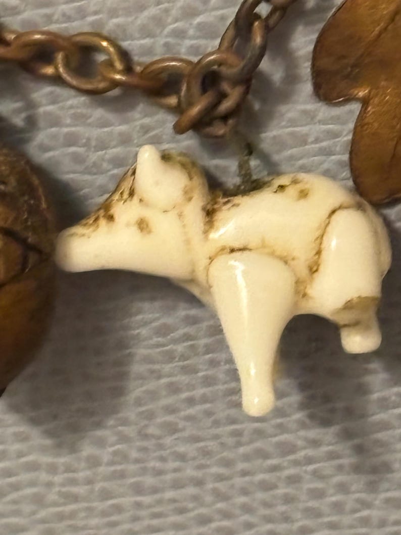 May include: A small, off-white pig charm with brown accents hangs from a bronze-colored chain. The pig is detailed with visible legs and a rounded body. The charm is part of a larger piece of jewelry.