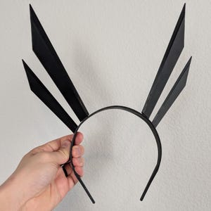 May include: A black headband with three upward-pointing, geometric shapes. The headband is held by a hand against a neutral background. The shapes are angled and create a striking, modern design. The headband is likely for fashion or costume use.