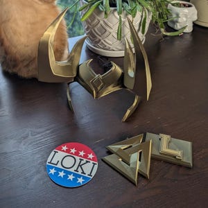 May include: A gold-colored Loki helmet with large horns, along with matching gold-colored accessories. A round sticker with the word "LOKI" in red, white, and blue is also present.