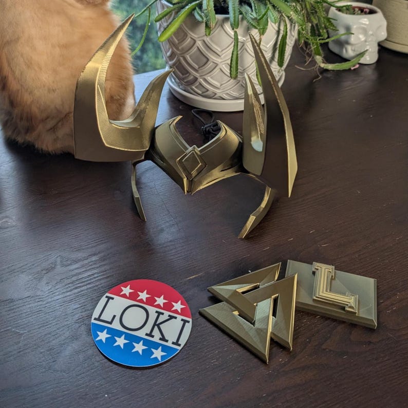 May include: A gold-colored Loki helmet with large horns, along with matching gold-colored accessories. A round sticker with the word "LOKI" in red, white, and blue is also present.