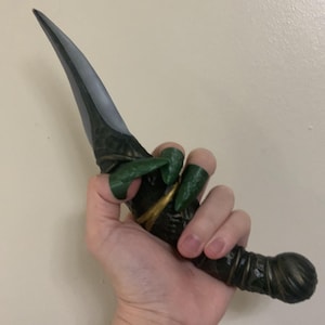 May include: A decorative dagger with a silver blade and a dark green and gold handle. The handle has a textured design. The dagger is held in a hand with green claw-like finger covers.