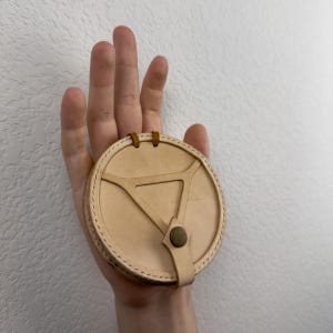 May include: A circular, light tan leather case featuring a triangular cutout design on the front surface. The case is secured with a small, dark olive green snap button closure and includes two small leather loops at the top edge for attachment.