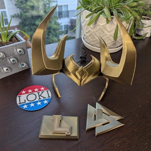 May include: A gold-colored Loki helmet with large horns, a circular "Loki" sticker, and two gold-colored geometric shapes. The helmet is the focal point, with the sticker and shapes arranged nearby on a dark surface.