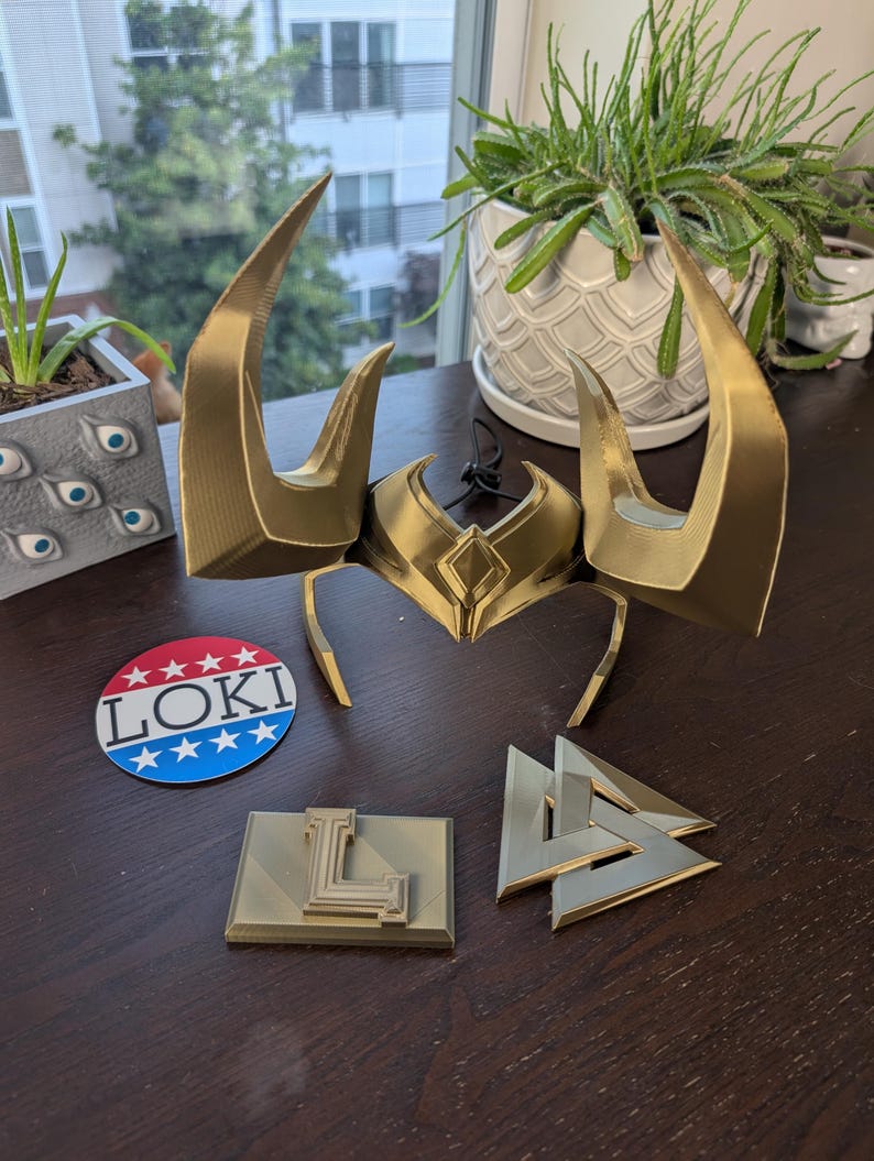 May include: A gold-colored Loki helmet with large horns, a circular "Loki" sticker, and two gold-colored geometric shapes. The helmet is the focal point, with the sticker and shapes arranged nearby on a dark surface.