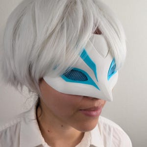 May include: A white fox-style mask featuring bright blue accents around the eye openings, which are covered with a blue mesh screen. The mask is worn over the upper face, partially obscured by a short, straight, white wig with layered bangs.