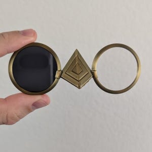 May include: Antique brass-colored glasses with a unique design. The glasses feature a circular lens, a triangular center piece, and a circular frame. The lens is dark, while the other parts are a warm brass color. The glasses are held by a hand.