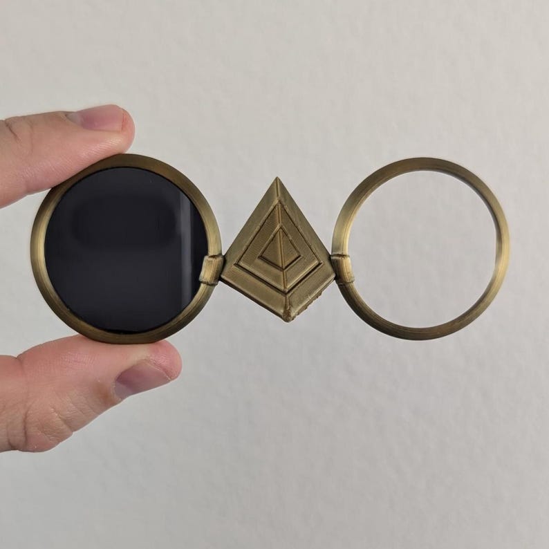 May include: Antique brass-colored glasses with a unique design. The glasses feature a circular lens, a triangular center piece, and a circular frame. The lens is dark, while the other parts are a warm brass color. The glasses are held by a hand.