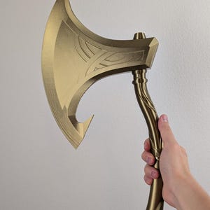 Puede incluir: Un hacha dorada con una hoja curva y un mango detallado. La cabeza del hacha presenta patrones decorativos grabados. El mango es sostenido por una mano, mostrando el tamaño y el diseño del hacha. Es un accesorio o un objeto decorativo.
