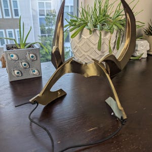 May include: A gold-colored, horned headpiece with a black adjustable cord. The headpiece has two large, curved horns and a central band. The horns are a darker shade of gold, and the piece appears to be made of metal or a similar material.