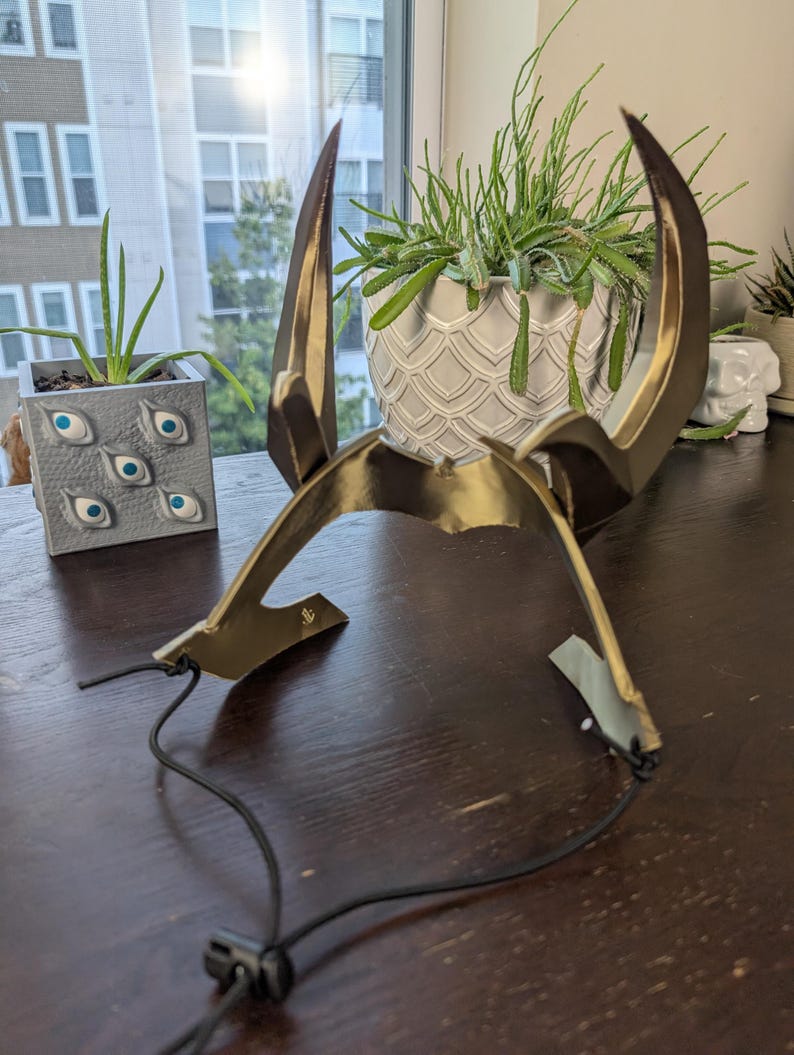 May include: A gold-colored, horned headpiece with a black adjustable cord. The headpiece has two large, curved horns and a central band. The horns are a darker shade of gold, and the piece appears to be made of metal or a similar material.