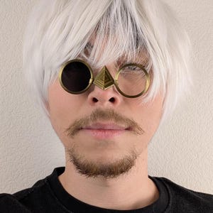 May include: A close-up of a person wearing unique steampunk-style sunglasses. The glasses feature a round, dark lens on the left, a clear circular frame on the right, and a gold pyramid-shaped bridge. The person has a mustache and beard.