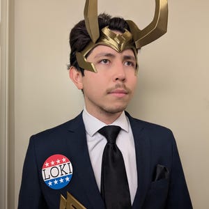 May include: A person wearing a gold-colored Loki helmet with large horns, a navy blue suit, and a black tie. A button that says "LOKI" is pinned to the suit. A gold-colored symbol is also pinned to the suit.