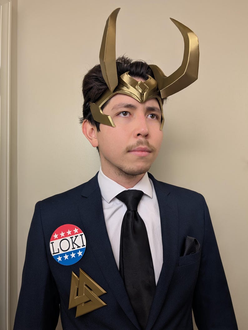 May include: A person wearing a gold-colored Loki helmet with large horns, a navy blue suit, and a black tie. A button that says "LOKI" is pinned to the suit. A gold-colored symbol is also pinned to the suit.