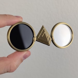 May include: A pair of round, antique brass-colored glasses with a dark lens on the left and a reflective lens on the right. A triangular piece connects the two lenses.