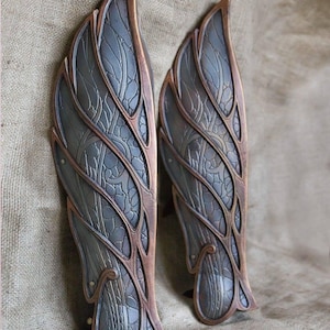 May include: A pair of decorative metal leg guards with a wing-like design. The guards are made of dark metal with a copper-colored trim and intricate patterns.
